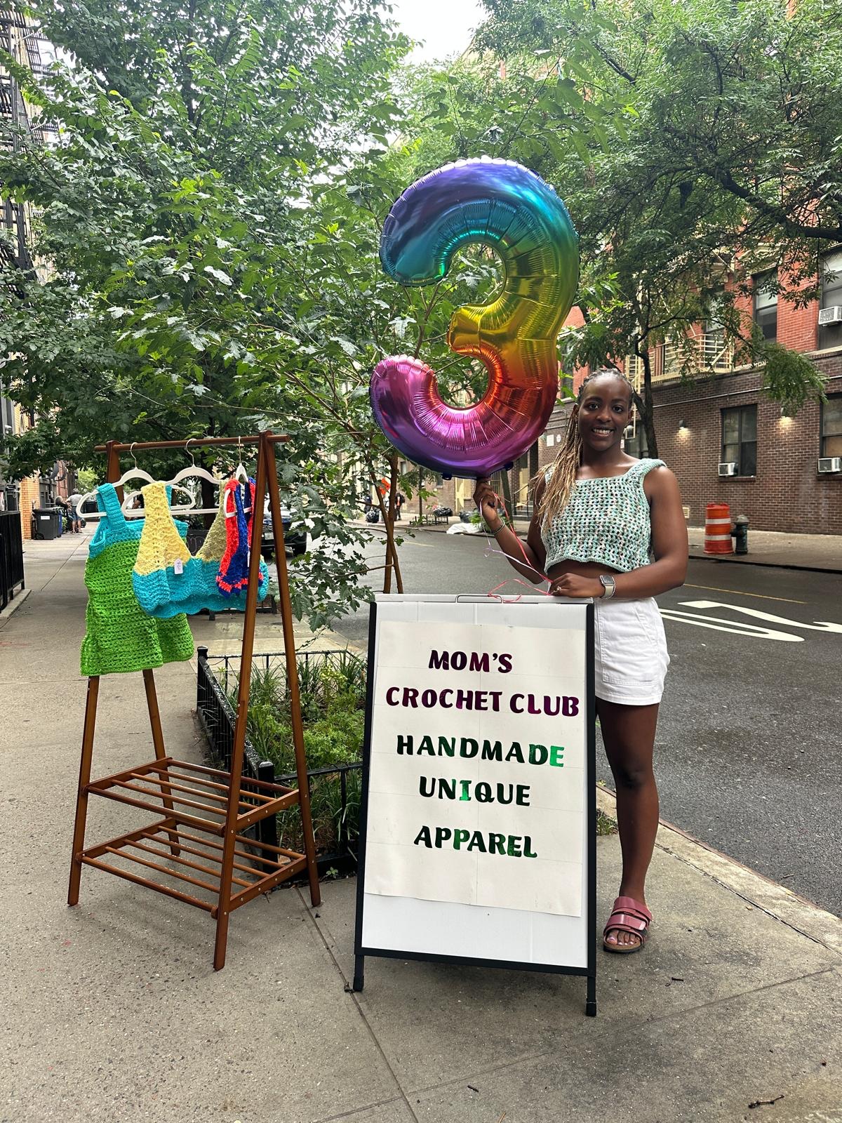 Mom's Crochet Club founder at a neighborhood market with handmade crochet apparel and a rainbow number three balloon
